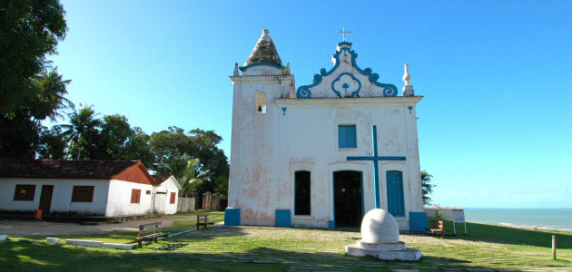 Igreja de Nossa Senhora da Conceição Barroca, construída no século XVII, no Centro Histórico de Santa Cruz Cabrália, com fachada branca, detalhes azuis e vista para o mar ao fundo.