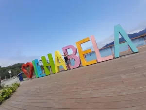 Letreiro colorido de Ilhabela em um deck de madeira à beira-mar, com uma pessoa sorrindo ao centro e montanhas ao fundo. Foto: Arquivo pessoal.