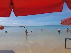 Turistas aproveitando o mar calmo e raso da Praia da Coroa Vermelha, em Santa Cruz Cabrália, na Bahia, em um dia de sol e céu azul, com barcos ao fundo e guarda-sóis na areia.