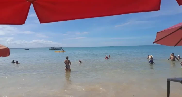 Turistas aproveitando o mar calmo e raso da Praia da Coroa Vermelha, em Santa Cruz Cabrália, na Bahia, em um dia de sol e céu azul, com barcos ao fundo e guarda-sóis na areia.