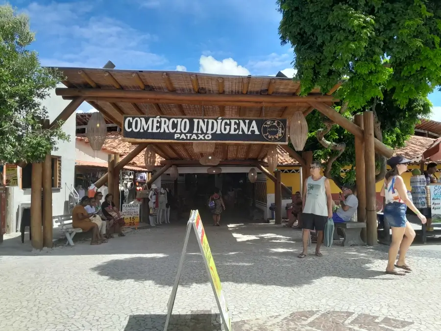 Vista da entrada do Comércio Indígena Pataxó, com alguns turistas sentados e outros andando em frente.
