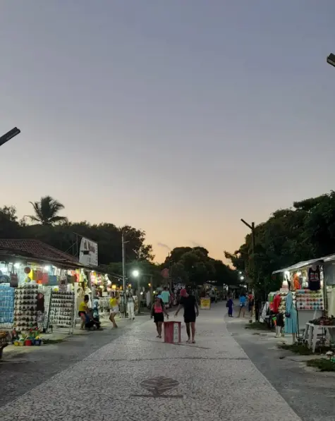 Vista da Feirinha de Coroa Vermelha ao entardecer, com turistas andando entre as barracas de roupas e artesanatos e belas árvores ao fundo.