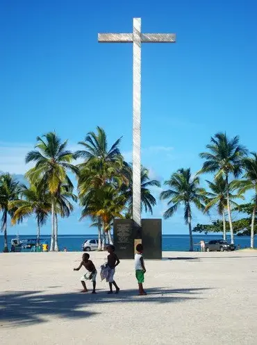 Marco simbólico da Primeira Missa no Brasil, na Praia da Coroa Vermelha, em Santa Cruz Cabrália, com uma cruz metálica, coqueiros ao fundo e crianças brincando no local.