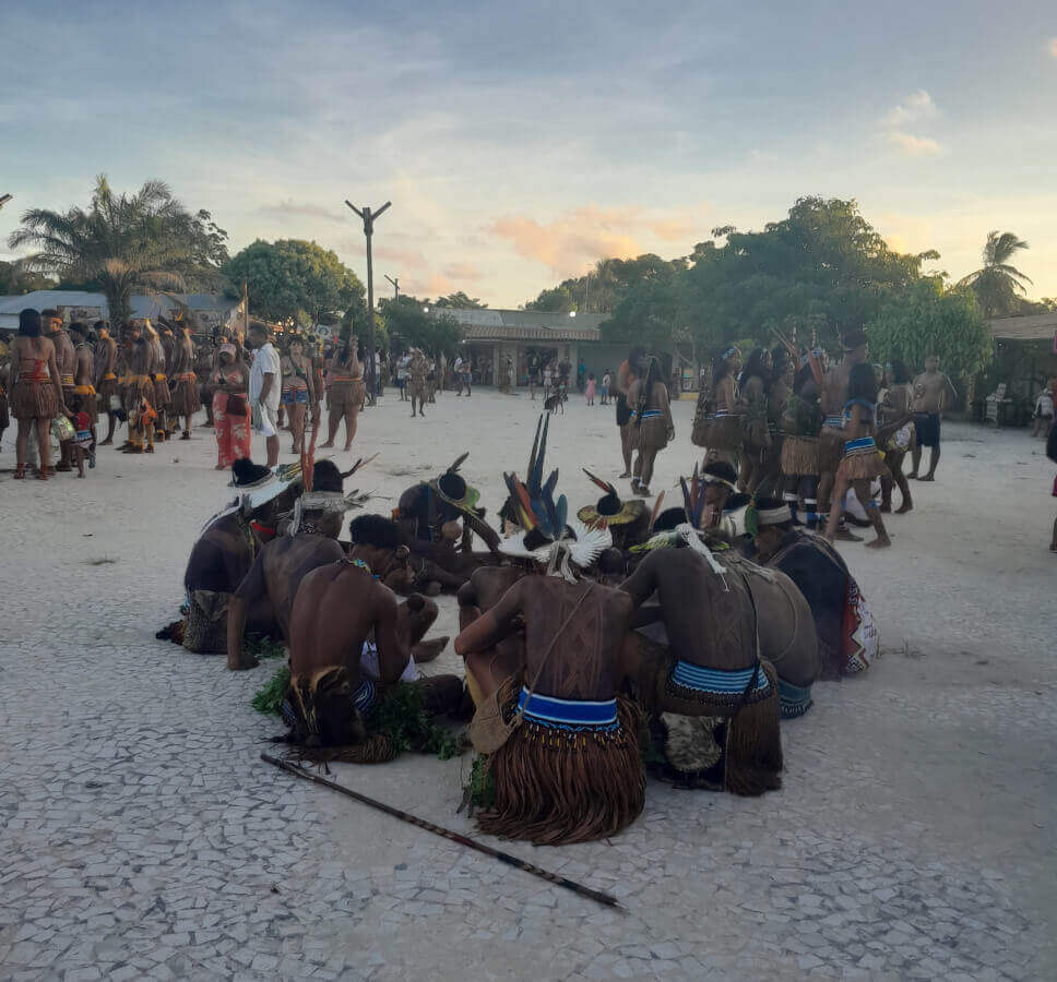 Indígenas Pataxó reunidos em roda, durante os Jogos Indígenas Pataxó em Santa Cruz Cabrália, na Bahia. Ao fundo, outras pessoas participam das atividades tradicionais.
