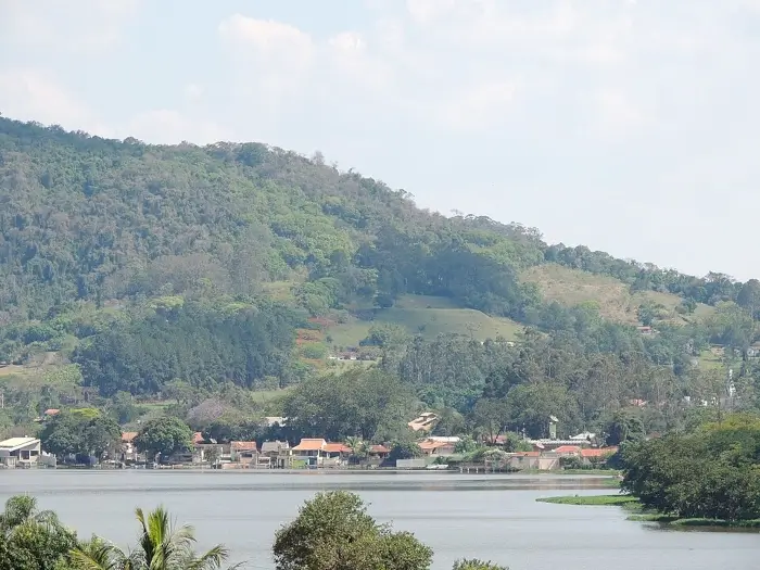 Vista da Represa Usina de Atibaia, com casas do outro lado da represa e uma grande montanha repleta de árvores atrás.