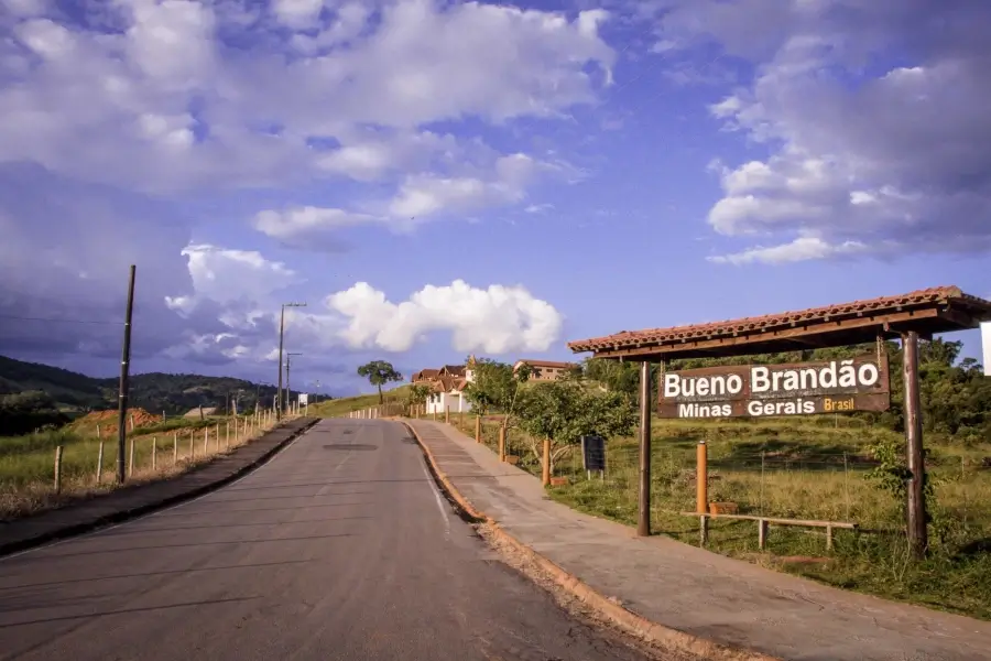 Placa de boas-vindas na entrada de Bueno Brandão, Minas Gerais, com a paisagem de uma estrada rural e montanhas ao fundo, sob um céu com nuvens.
