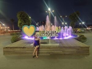 Marcia Lira, redatora e fundadora do Viagens e Passeios sentada em uma plataforma de madeira em frente a uma fonte luminosa colorida com a frase "SEU, MEU, NOSSO PORTO SEGURO" e um grande coração amarelo, em uma praça iluminada à noite em Porto Seguro, Bahia. Foto: arquivo pessoal.