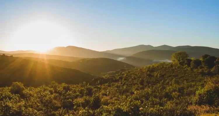Paisagem com montanhas verdes e o nascer do sol ao fundo, representando a região da Serra da Mantiqueira em Minas Gerais.