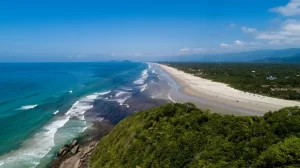 Vista aérea da Praia de Guaratuba em Bertioga, mostrando a longa e ampla faixa de areia branca, o mar com tons de azul e turquesa, e a extensa vegetação verde da restinga que a margeia, com as montanhas da Serra do Mar ao fundo. Foto: Prefeitura de Bertioga.
