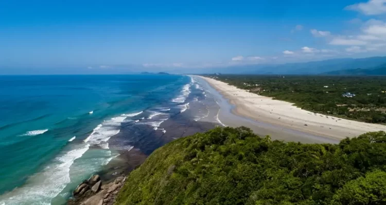 Vista aérea da Praia de Guaratuba em Bertioga, mostrando a longa e ampla faixa de areia branca, o mar com tons de azul e turquesa, e a extensa vegetação verde da restinga que a margeia, com as montanhas da Serra do Mar ao fundo. Foto: Prefeitura de Bertioga.