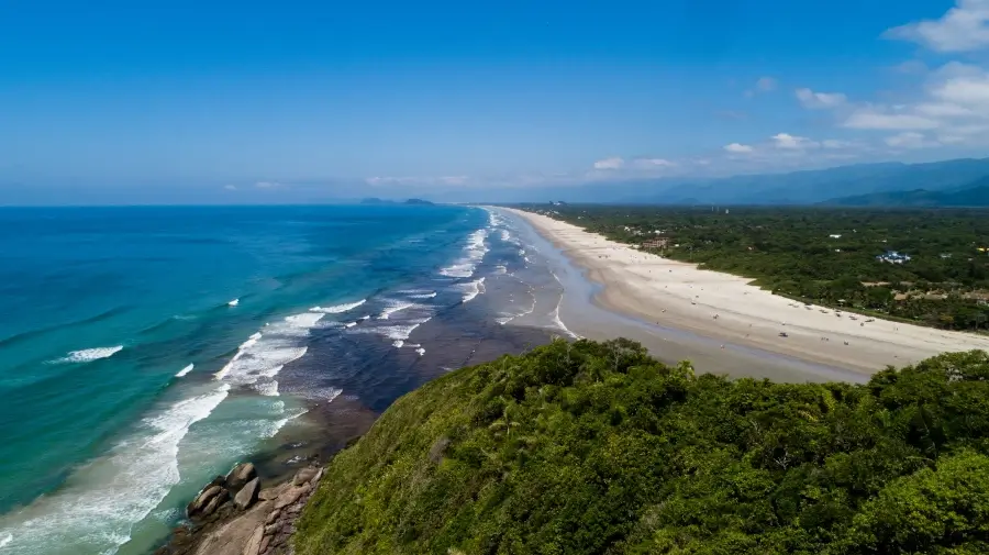 Vista aérea da Praia de Guaratuba em Bertioga, mostrando a longa e ampla faixa de areia branca, o mar com tons de azul e turquesa, e a extensa vegetação verde da restinga que a margeia, com as montanhas da Serra do Mar ao fundo. Foto: Prefeitura de Bertioga.
