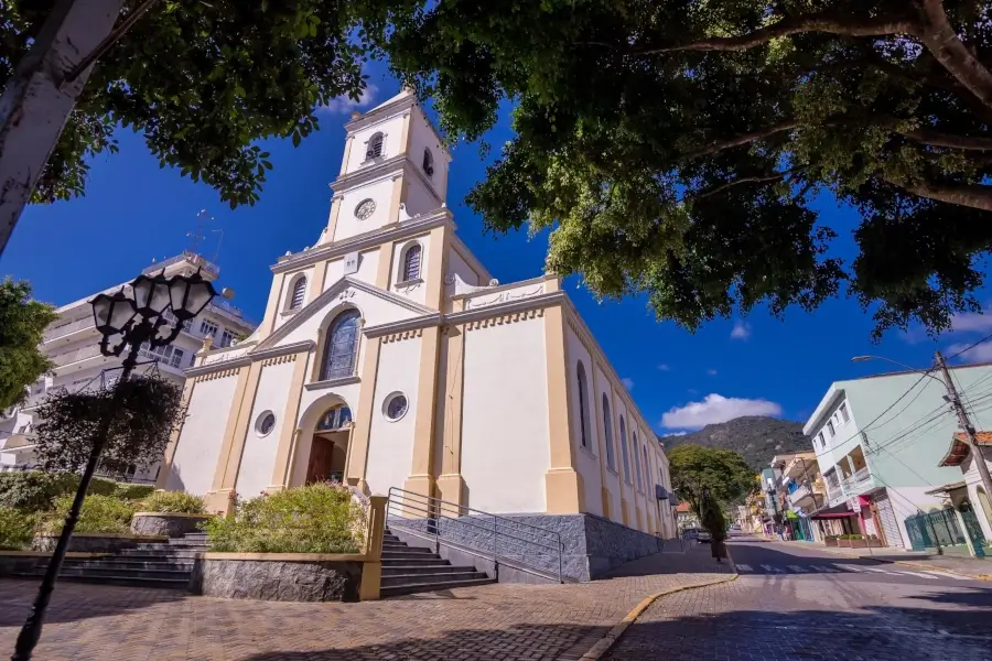 Fachada da Igreja Matriz de Extrema, Minas Gerais, uma construção branca e amarela com uma torre alta, vista de baixo com árvores em primeiro plano e o céu azul com nuvens.