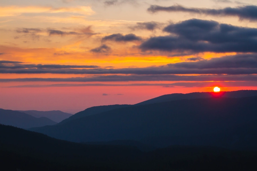 Cena do pôr do sol nas montanhas representando a Rota do Sol em Extrema, Minas Gerais, com o sol se pondo atrás das montanhas em um céu com tons de laranja, rosa e roxo, e nuvens escuras.