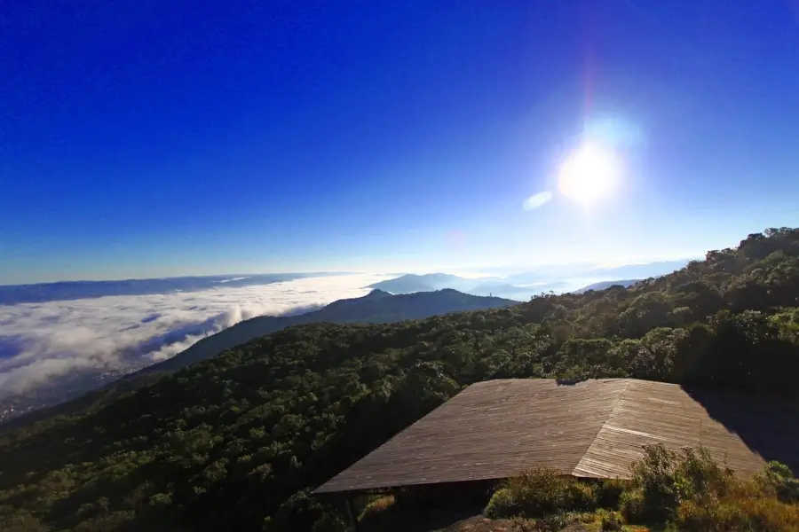 Vista panorâmica do alto da Rota dos Ventos em Extrema, Minas Gerais, com montanhas verdejantes, um mar de nuvens no horizonte e o céu azul-claro. Uma plataforma de madeira é visível em primeiro plano.