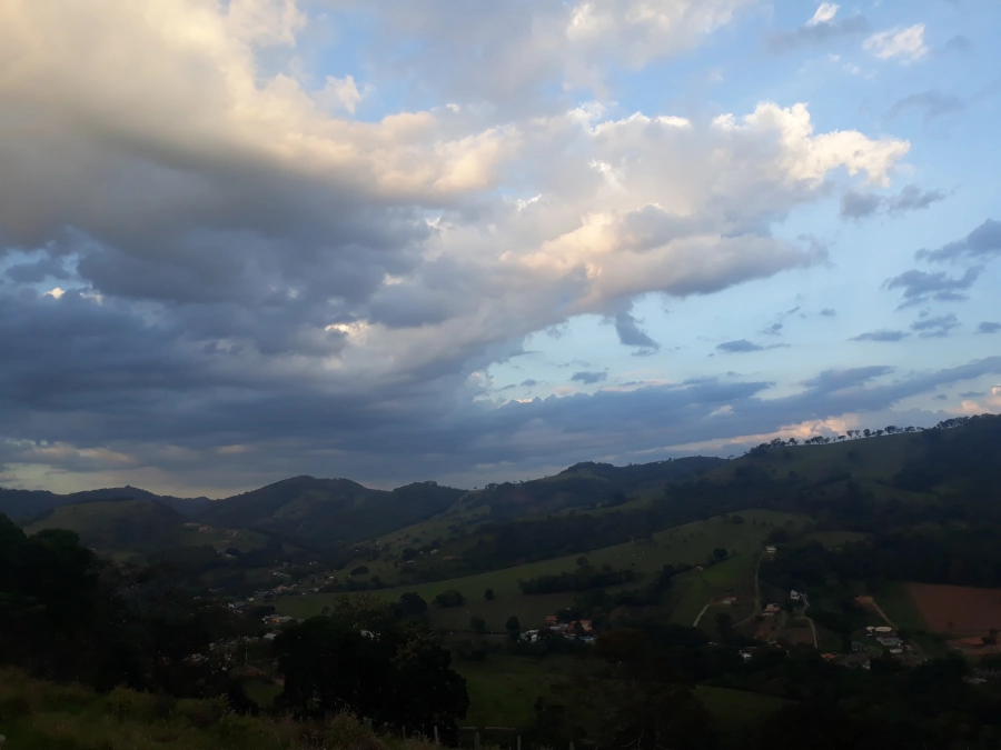 Ampla vista panorâmica das montanhas verdejantes e vales de Extrema, Minas Gerais, sob um céu com diversas camadas de nuvens em tons de azul e cinza.