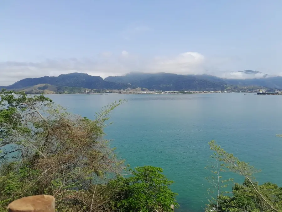 Vista de um dos mirantes de Ilhabela, com algumas árvores nativas à frente, o mar verde no centro e as montanhas com nuvens atrás.