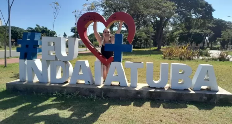 Letreiro "Eu amo Indaiatuba" com a Marcia Lira, redatora do blog Viagens e Passeios, atrás dele. Há árvores ao fundo mostrando que a cidade é bem arborizada.