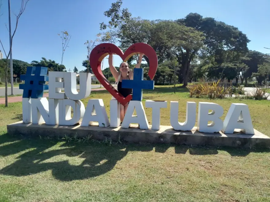 Letreiro "Eu amo Indaiatuba" com a Marcia Lira, redatora do blog Viagens e Passeios, atrás dele. Há árvores ao fundo mostrando que a cidade é bem arborizada.