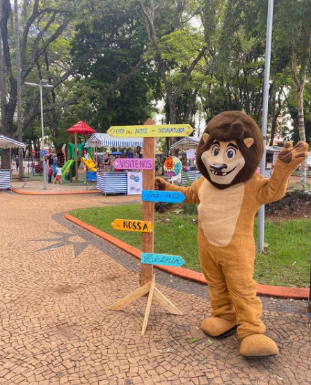 Mascote vestido de leão posa ao lado de placa colorida durante a Feira de Artes de Indaiatuba, com barracas e parquinho ao fundo.