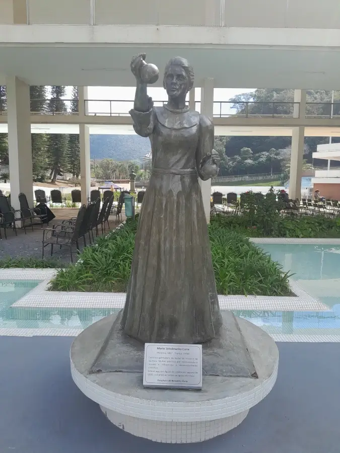 Estátua de bronze de Marie Curie em Águas de Lindóia, segurando um objeto, com uma placa de identificação na base.