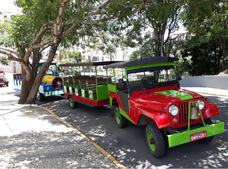Um pequeno trem turístico de cores vibrantes, com uma locomotiva em forma de jipe vermelho e verde, parado na rua.