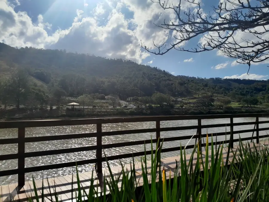 A Represa Cavalinho Branco em Águas de Lindóia, com a água calma, uma cerca de madeira, e montanhas no horizonte em um dia ensolarado.