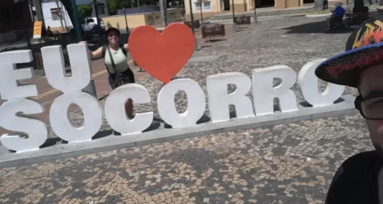 Casal do Viagens e Passeios - Marcia Lira (fundadora e redatora) e Windsor (fundador e analista SEO) - tira selfie em frente ao letreiro “Eu amo Socorro” na praça da matriz da cidade, com a igreja ao fundo.
