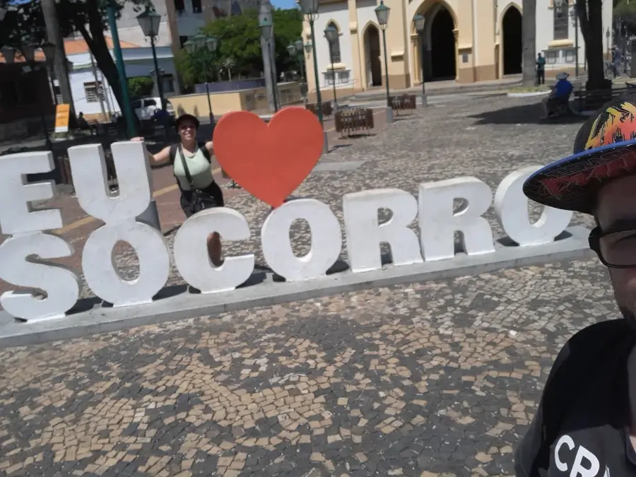 Casal do Viagens e Passeios - Marcia Lira (fundadora e redatora) e Windsor (fundador e analista SEO) - tira selfie em frente ao letreiro “Eu amo Socorro” na praça da matriz da cidade, com a igreja ao fundo.