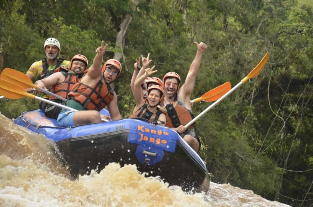 Grupo de pessoas com capacetes e coletes salva-vidas em um bote de rafting azul, remando e celebrando enquanto descem uma corredeira em rio de águas barrentas, com floresta ao fundo.