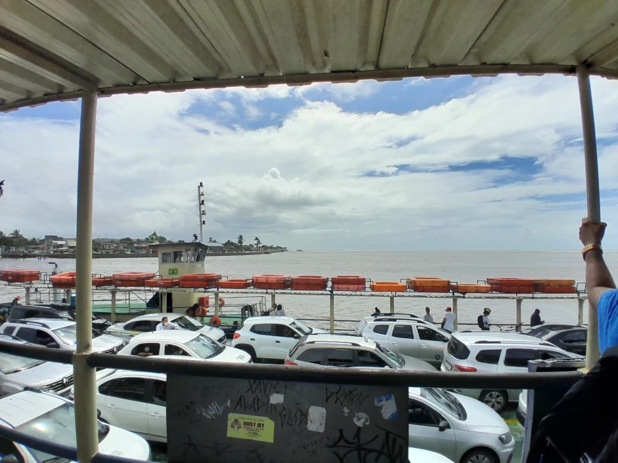Vista do interior da balsa que faz a travessia do Rio Buranhém, com o deck inferior repleto de carros e, ao fundo, a orla de Porto Seguro vista da água.