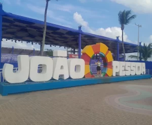 Marcia Lira, fundadora e redatora do Viagens e Passeios, sorrindo e posando ao lado do letreiro “JOÃO PESSOA” na orla da cidade, com o mar ao fundo e coqueiros, representando o que fazer em João Pessoa em 7 dias.
