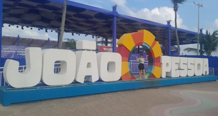 Marcia Lira, fundadora e redatora do Viagens e Passeios, sorrindo e posando ao lado do letreiro “JOÃO PESSOA” na orla da cidade, com o mar ao fundo e coqueiros, representando o que fazer em João Pessoa em 7 dias.