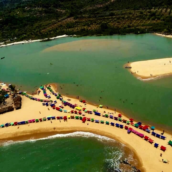 Vista aérea da ponta de areia em Caraíva, Bahia, mostrando o encontro do Rio Caraíva, de águas esverdeadas, com o mar. A praia está repleta de guarda-sóis coloridos e banhistas.