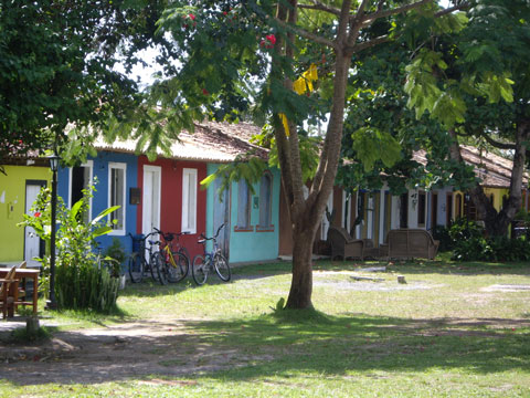 Fileira de casas coloniais coloridas no Quadrado de Trancoso, Bahia, com bicicletas estacionadas em frente, sob a sombra de árvores em um gramado.