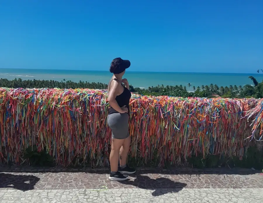 Marcia Lira, redatora do Viagens e Passeios, em pé no Mirante das Fitas em Arraial d'Ajuda, com o mar azul e a linha de coqueiros ao fundo, contemplando a paisagem.