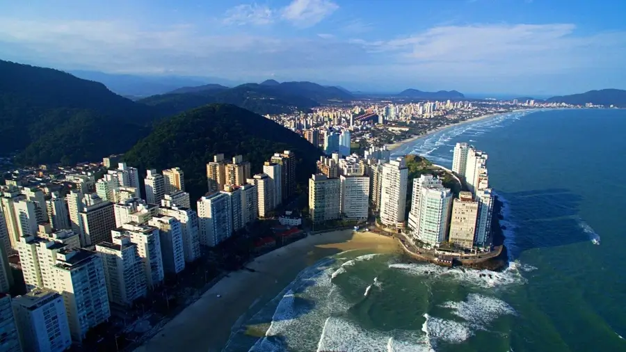 Vista aérea panorâmica da orla urbana de Guarujá, destacando a alta densidade de prédios frente ao mar na Praia de Pitangueiras e as montanhas verdes ao fundo.