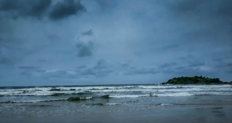 Paisagem de uma praia em Guarujá sob céu nublado e dramático, com ondas brancas quebrando no mar escuro e um morro com vegetação ao fundo à direita. Foto: arquivo pessoal, melhorada com Gemini.
