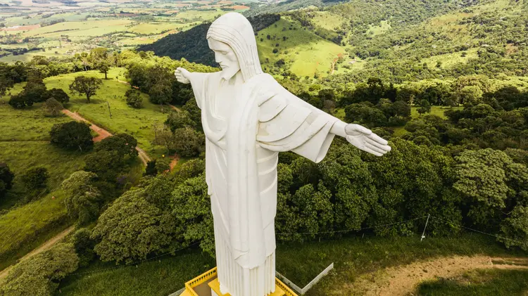 Vista aérea do monumento Cristo Redentor de Pouso Alegre, estátua branca de braços abertos sobre uma montanha verde.
