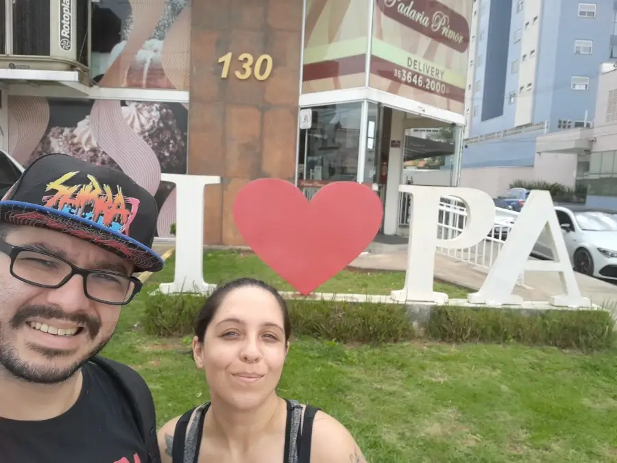 Casal de viajantes fundadores do Viagens e Passeios, Marcia Lira e Windsor Fernandes, tirando uma selfie sorrindo em frente ao letreiro turístico com um coração vermelho e as letras PA.