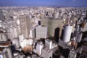 Vista aérea panorâmica da densa cidade de São Paulo, destacando edifícios emblemáticos do centro como o Copan e o Edifício Itália sob um céu azul claro, mostrando a imensidão urbana. Foto: Prefeitura de São Paulo.