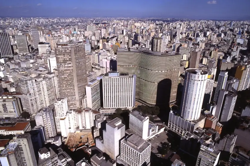 Vista aérea panorâmica da densa cidade de São Paulo, destacando edifícios emblemáticos do centro como o Copan e o Edifício Itália sob um céu azul claro, mostrando a imensidão urbana. Foto: Prefeitura de São Paulo.