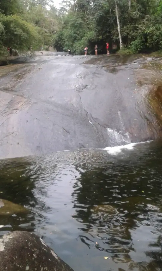 Fotografia em ângulo baixo mostrando a base da Cachoeira do Tobogã em Paraty, RJ. A água escura da piscina natural está em primeiro plano, refletindo a floresta. Ao fundo, uma grande e lisa laje de pedra escura por onde a água escorre. No topo da pedra, algumas pessoas estão em pé, observando a queda d'água, cercadas por uma vegetação densa e verde de Mata Atlântica. O céu está encoberto e cinzento.