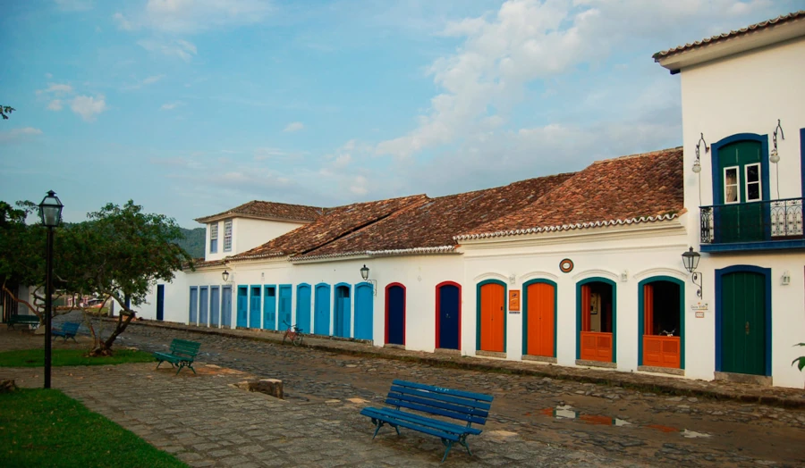 Fotografia ampla de uma rua de pedra "pé de moleque" no Centro Histórico de Paraty em um dia ensolarado. Em primeiro plano, uma praça com bancos de madeira pintados de azul e um poste de luz antigo em estilo colonial. Ao fundo, uma fileira de casarões térreos brancos com portas e janelas coloridas em tons de azul, vermelho e amarelo. O céu está azul claro com poucas nuvens.