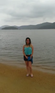Marcia Lira, redatora e fundadora do Viagens e Passeios, em pé, de corpo inteiro, posando em uma praia de areia clara em Paraty, RJ. Ela é uma mulher com cabelos pretos e ombreiras loiras, usando uma blusa turquesa de alças e shorts pretos. Ela segura uma canga azul e sorri levemente para a câmera. Ao fundo, a Baía da Ilha Grande em um dia com céu nublado e cinzento, e morros cobertos de verde no horizonte.