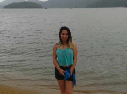 Marcia Lira, redatora e fundadora do Viagens e Passeios, em pé, de corpo inteiro, posando em uma praia de areia clara em Paraty, RJ. Ela é uma mulher com cabelos pretos e ombreiras loiras, usando uma blusa turquesa de alças e shorts pretos. Ela segura uma canga azul e sorri levemente para a câmera. Ao fundo, a Baía da Ilha Grande em um dia com céu nublado e cinzento, e morros cobertos de verde no horizonte.