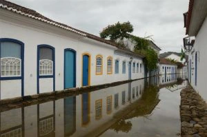 Vista de uma rua estreita no Centro Histórico de Paraty durante o fenômeno da maré alta. A água do mar cobre completamente o calçamento de pedras, criando um espelho perfeito que reflete as fachadas brancas dos casarões e as molduras coloridas das portas. O céu está nublado e o reflexo na água acentua a arquitetura simétrica da cidade.