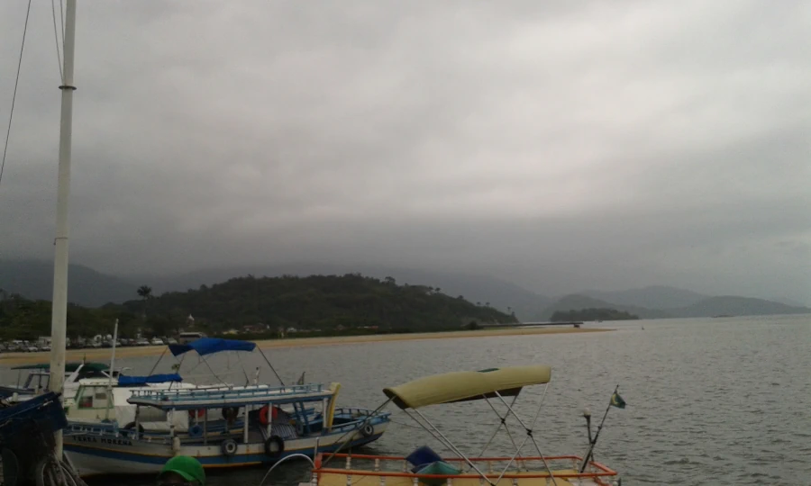 Vista panorâmica do Cais de Paraty em um dia com céu totalmente encoberto por nuvens cinzentas e densas. No primeiro plano, à esquerda, barcos de pesca e escunas de turismo coloridas estão atracados na água calma e escura. Uma longa faixa de areia clara separa a água de morros cobertos por vegetação verde escura ao fundo, que se estendem até o horizonte sob o céu pesado de chuva.