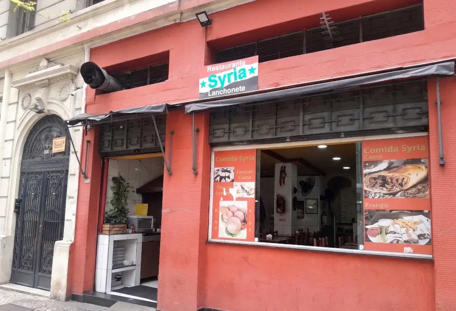 Fachada do restaurante Syria, no centro de São Paulo. A fachada é vermelha, com banners com fotos de comidas típicas árabes nas janelas, e a placa com o nome do restaurante na parte superior.