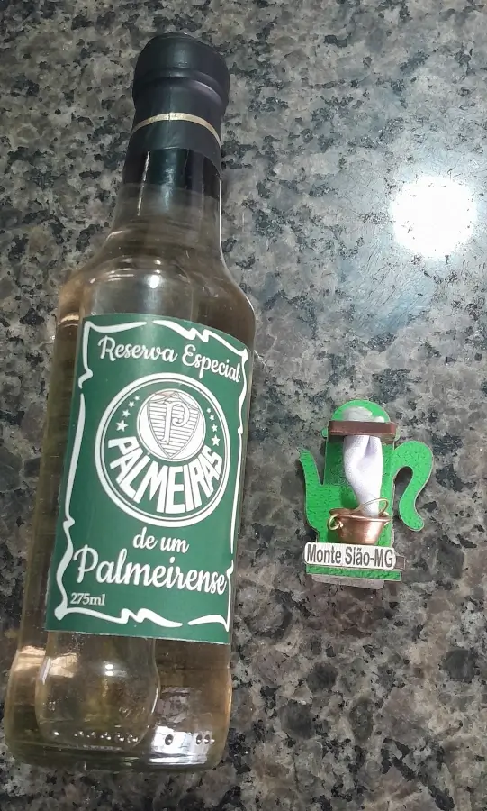 Uma garrafa pequena de cachaça artesanal com rótulo verde do Palmeiras ao lado de um ímã de geladeira artesanal em formato de bule com a inscrição Monte Sião-MG.