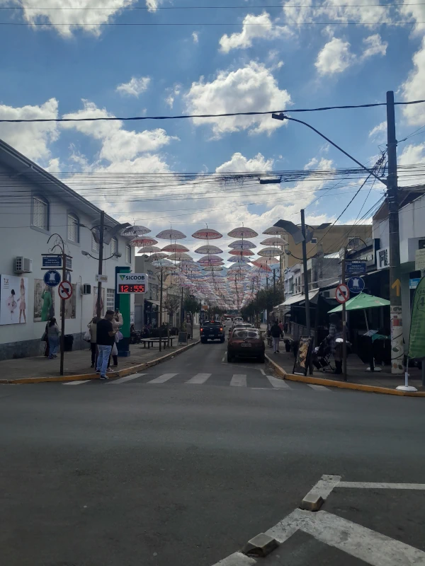 Rua comercial no centro de Monte Sião decorada com dezenas de guarda-chuvas coloridos suspensos no céu sob luz solar.